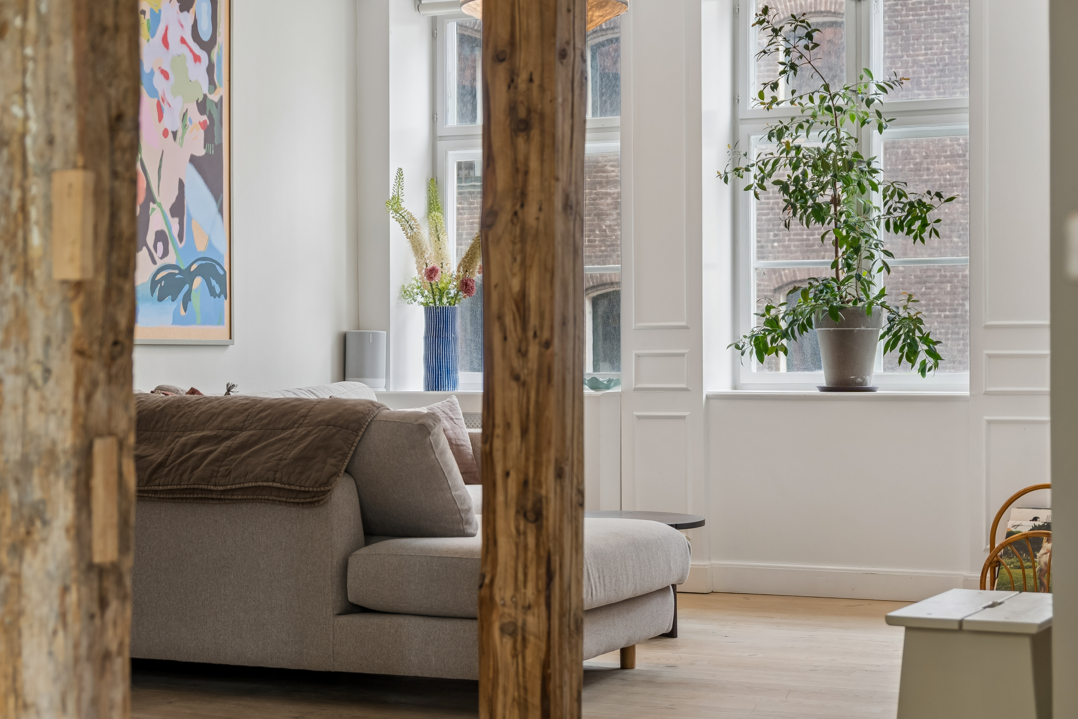 Stue med grå sofa, synlige træstolper, et farverigt billede på væggen og planter i vindueskarmene.