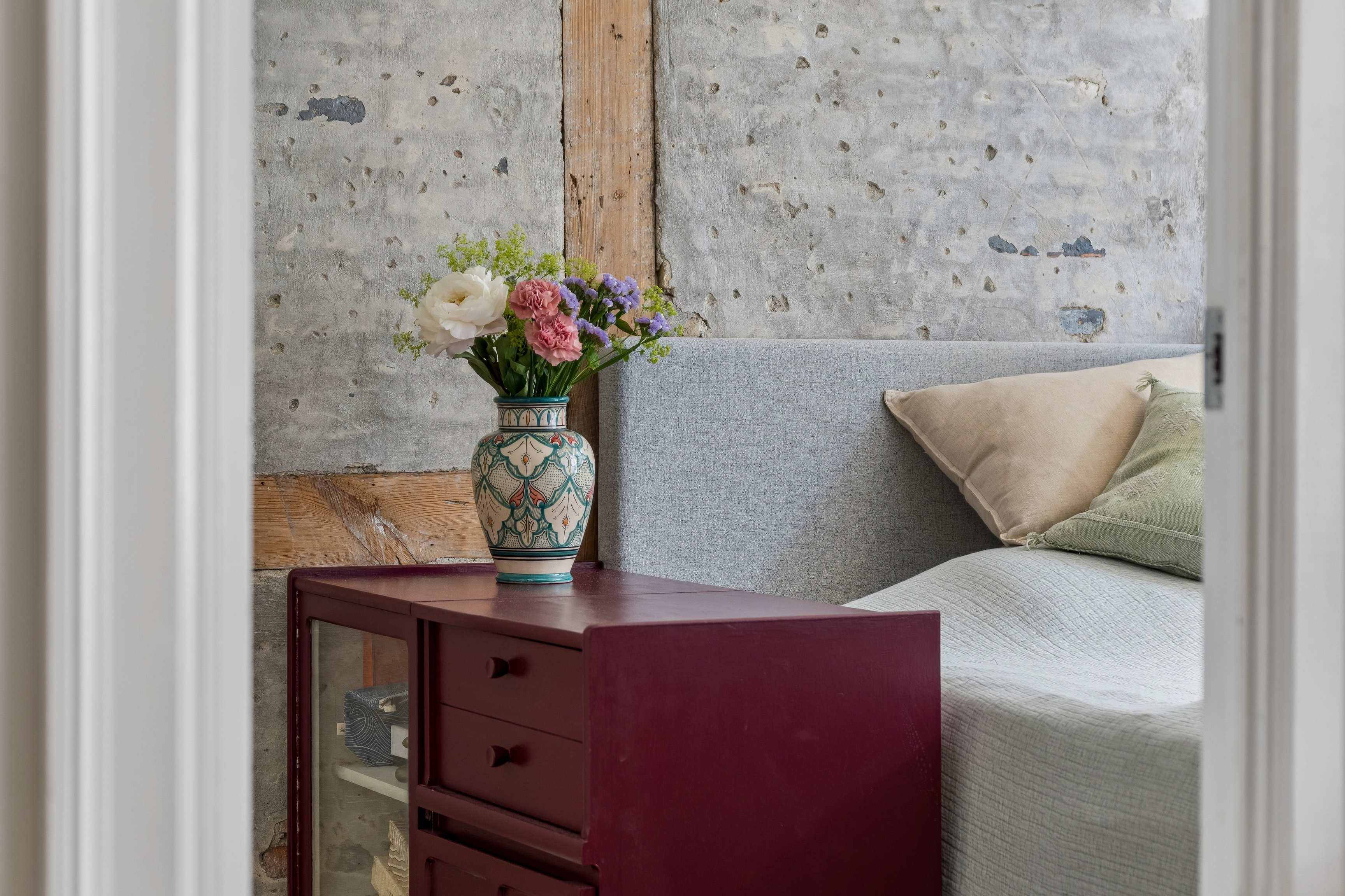 En mønstret vase med blomster står på et vinrødt skab ved siden af en seng med gråt sengetæppe og beige og grønne puder foran en rå væg med synligt træværk.