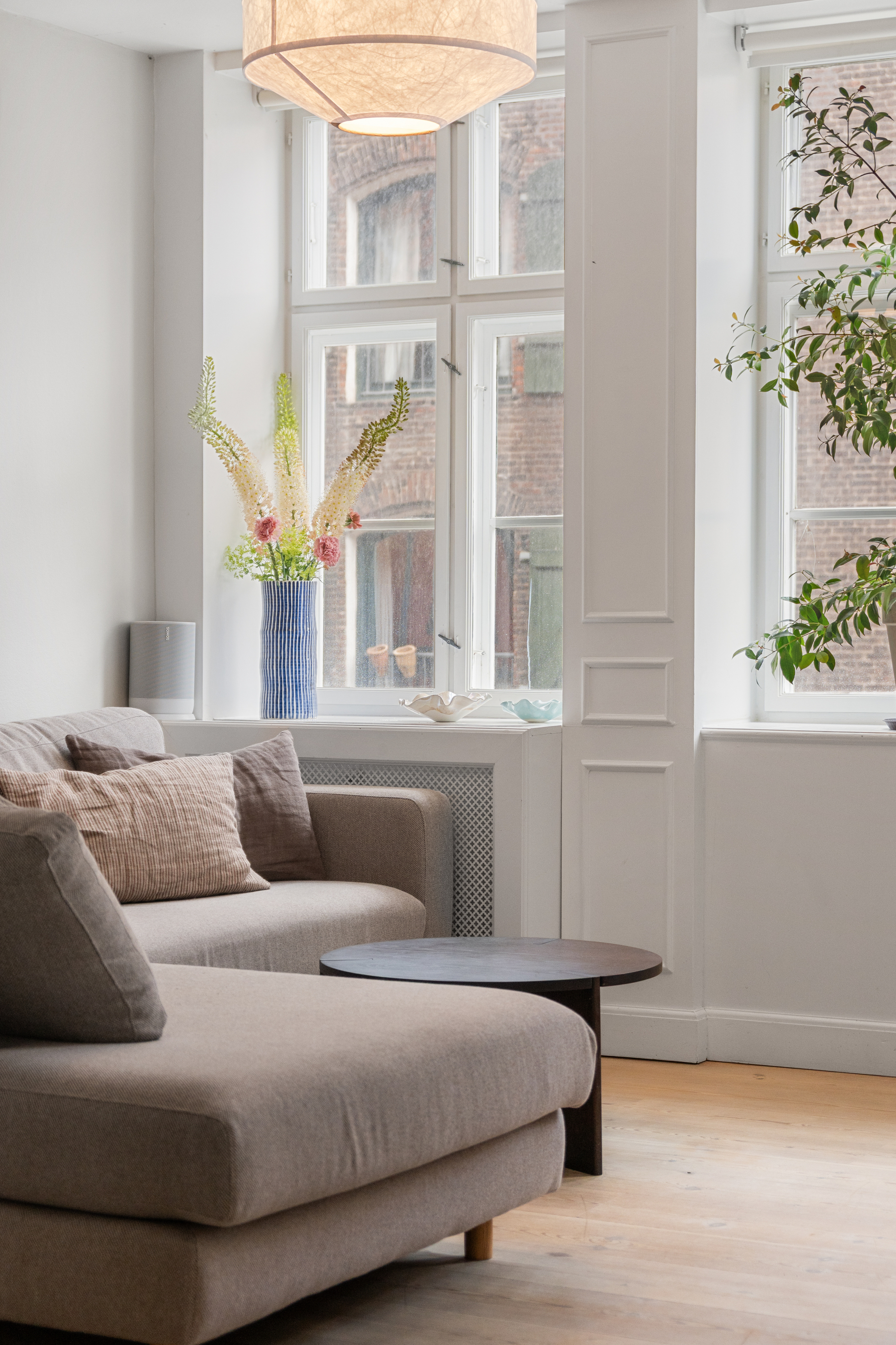 Stue med grå sofa med chaiselong, et mørkt rundt sofabord, en stor pendel og blomster i en stribet vase foran høje vinduer.
