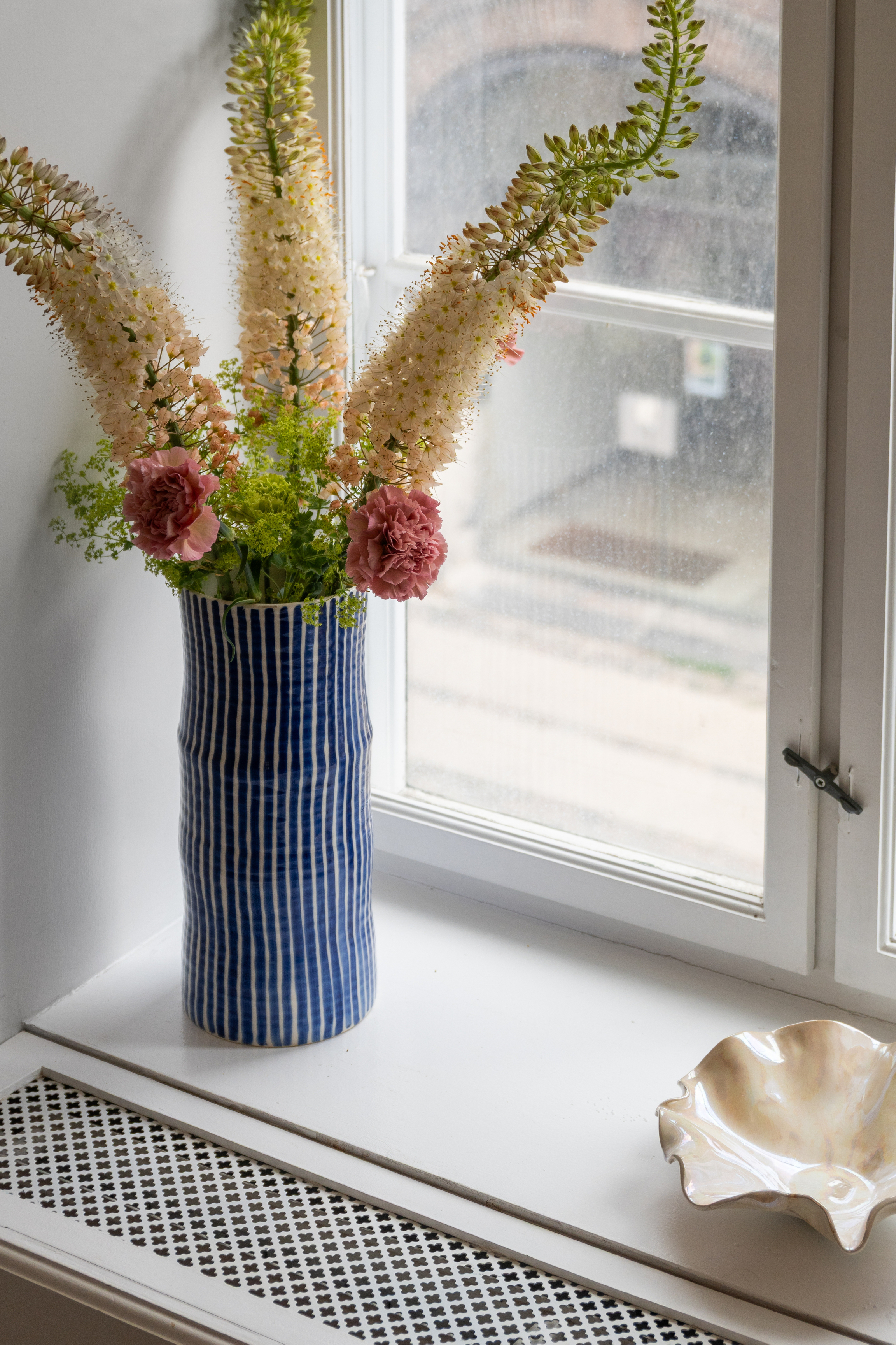 En blå- og hvidstribet vase med høje lyse blomster, lyserøde nelliker og grønt står på en hvid vindueskarm ved siden af en skål med bølget kant.