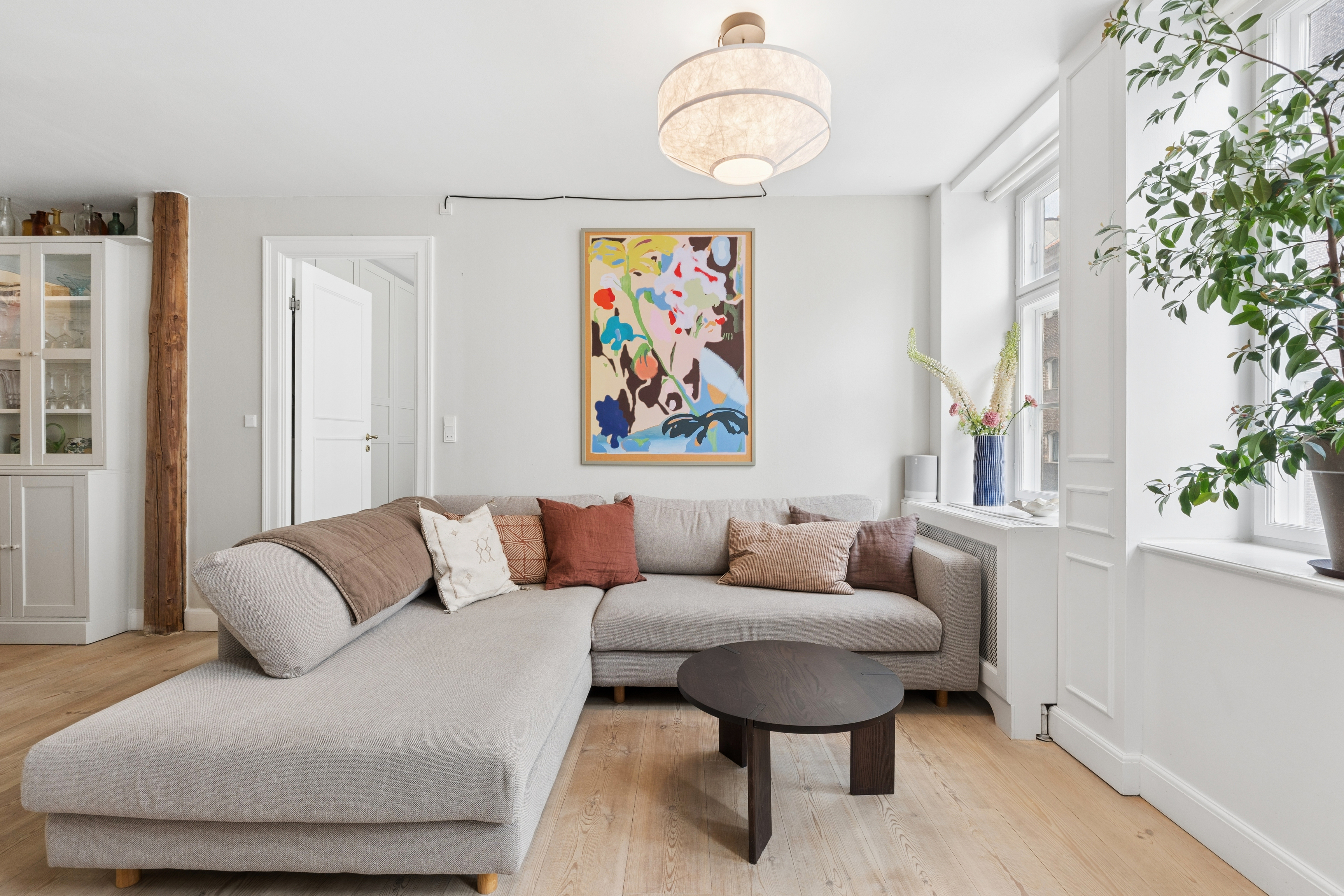 Stue med en lysegrå hjørnesofa, et mørkt sofabord, et farverigt maleri og store vinduer med planter og blomster.