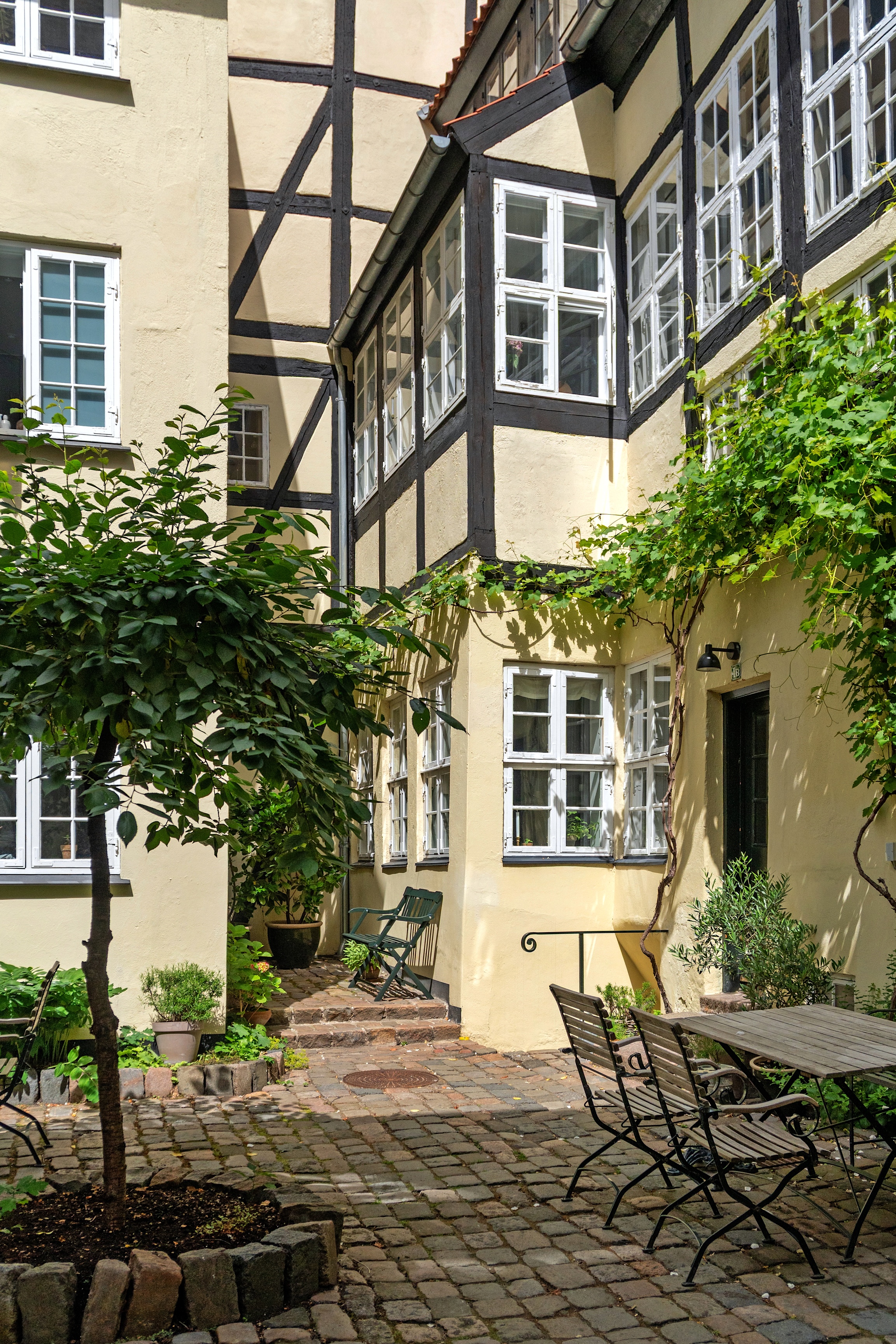 En brostensbelagt gårdhave i Christianshavn med havemøbler, et træ og klatreplanter foran en gul bindingsværksbygning med hvide vinduer.